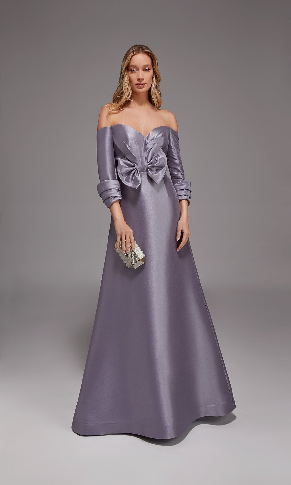 A woman in the Alyce 27859 long formal dress, featuring an off-the-shoulder lilac grey satin design with a bow at the waist and a silver clutch, stands against a gray background—a perfect look for mothers of the bride or anyone seeking elegance.